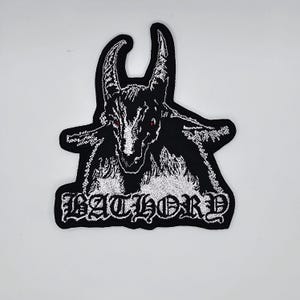 May include: Black and white embroidered patch featuring a goat-like creature with red eyes. The patch has the word "BATHORY" in a gothic font at the bottom. The patch is likely for a jacket or bag.