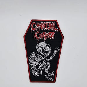 May include: Black coffin-shaped patch with a red border and the band name "Cannibal Corpse" in red, dripping font. A white skeleton is depicted inside the coffin, with its arms raised. The patch is likely for a jacket or bag.