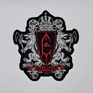 Peut inclure: Écusson noir brodé avec un motif de crête rouge et noir. Il présente une couronne, des lions et un emblème stylisé, avec le mot "EMPEROR" sur une bannière rouge.