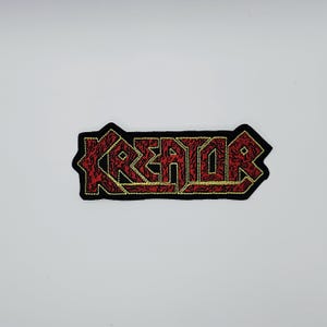 Może przedstawiać: Haftowana naszywka z napisem "KREATOR" w kolorze czerwonym, obramowana złotymi przeszyciami na czarnym tle. Naszywka ma charakterystyczną, postrzępioną obwódkę. Tekst jest pogrubioną, stylizowaną czcionką.