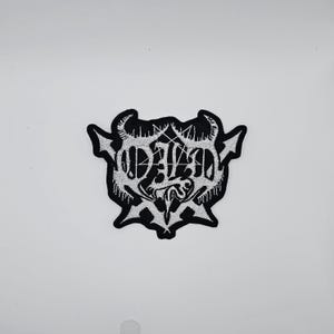 Puede incluir: Parche bordado en blanco y negro con la palabra "DALAD" en una fuente gótica, con un pentagrama y elementos decorativos. El parche tiene un aspecto texturizado y un diseño único y estilizado.