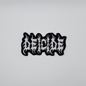 May include: Black and white embroidered patch with the word "DEICIDE" in a gothic font. The patch has a black border and the text is in white. The patch is rectangular in shape.