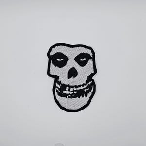 Puede incluir: Parche bordado con el icónico logotipo de la calavera de Misfits. El parche tiene un borde negro, con una calavera blanca y cuencas de ojos negras. La boca de la calavera está abierta, revelando dientes. Adecuado para chaquetas, bolsos u otros artículos de tela.