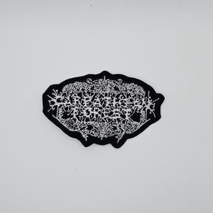 Pode incluir: Um patch bordado em preto e branco com o texto "REAPER CORPSE" em uma fonte gótica estilizada. O patch tem uma forma irregular e é projetado para aplicação em roupas ou acessórios.