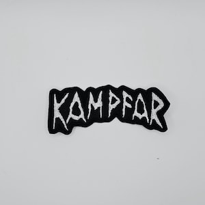 Può includere: Una toppa ricamata nera con la scritta "KAMPFAR" in bianco, con lettere irregolari. La toppa ha un bordo nero e una forma rettangolare. Lo sfondo è bianco.