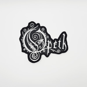May include: A black and white embroidered patch featuring the band name "Opeth" in a gothic font. The patch has a unique design with ornate swirls and a stylized letter "O". The patch is likely for clothing or accessories.