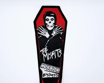 May include: Black and red patch featuring a coffin design with a skeletal figure and the text "The Misfits Horror Business". The patch has a black border and is likely for clothing or accessories.