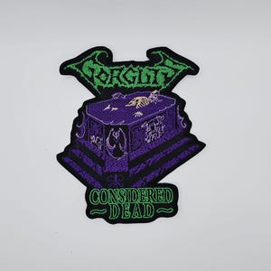 Puede incluir: Parche bordado con un diseño de ataúd morado y esqueleto, la palabra "Gorguts" en verde arriba y "Considered Dead" en verde abajo. El parche tiene un borde negro y está diseñado para ropa o bolsos.