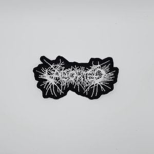 Może przedstawiać: Czarna, haftowana naszywka z napisem "ABORTED" w białych, stylizowanych, kolczastych literach. Naszywka ma unikalny, nieregularny kształt i jest prawdopodobnie przeznaczona do odzieży lub akcesoriów.