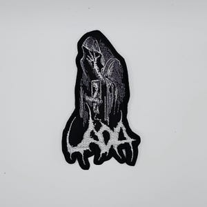 以下が含まれることがあります： 本を持っているフード付きの人物が描かれた、白黒の刺繍パッチ。人物は陰影とテクスチャで詳細に描かれており、パッチの下部には「GOAT」という言葉が様式化されたフォントで書かれています。