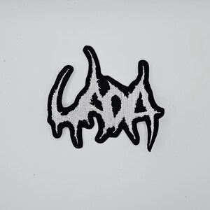 以下が含まれることがあります： スタイリッシュなドリップフォントで「SOA」という文字が刺繍された白黒のパッチ。パッチは黒い縁取りと白いテクスチャ加工された内側を備え、コントラストの強いデザインになっています。衣類やアクセサリーに最適です。