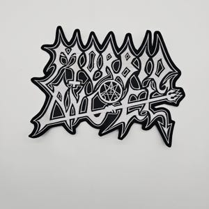 May include: A black and white embroidered patch with a jagged border. The patch features stylized text and symbols, including a pentagram and a trident. The design is intricate and detailed, with a heavy metal or gothic aesthetic.