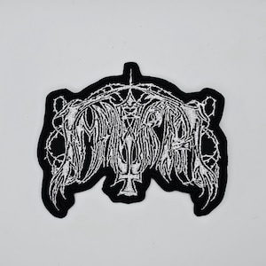 May include: Black and white embroidered patch with a gothic design. The patch features intricate white stitching on a black background, with a central emblem and ornate detailing. The design includes a cross and stylized text.