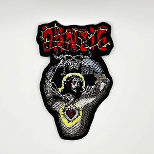May include: An embroidered patch featuring a black background with the red text "DENTIC" at the top. Below the text is a skull and a figure with a yellow halo surrounding a red heart, rendered in black, white, and yellow thread with detailed stitching.