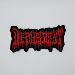 Könnte beinhalten: Ein schwarzer, gestickter Aufnäher mit dem Wort "DEVOURMENT" in roter, stilisierter, tropfender Schrift. Der Aufnäher hat einen gezackten, unregelmäßigen Rand. Der Text ist im Heavy-Metal-Stil gehalten.