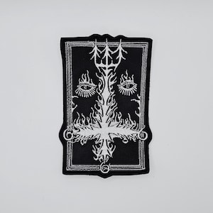 May include: Black and white embroidered patch featuring a stylized design. The patch has a rectangular shape with a decorative border. The central design includes a cross, flames, and eye symbols.