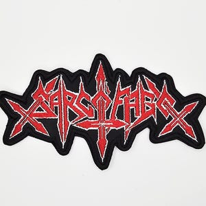 May include: Embroidered patch with the word "XARSCABEX" in red, stylized lettering. The patch has a black border and a white outline around the red text. The design features sharp, angular elements.
