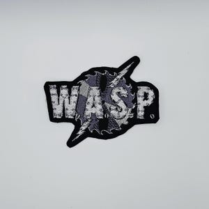 May include: Embroidered patch with the word "WASP" in white, stylized letters. The letters are set against a gray and black checkered background, with a lightning bolt design. The patch has a black border.