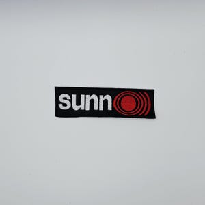 May include: Black rectangular patch with the word "sunn" in white, sans-serif font. A red concentric circle design is on the right side. The patch is likely for clothing or accessories.