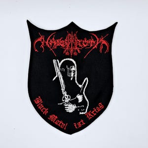 May include: Black shield-shaped patch with red and white embroidered design. The patch features a stylized figure holding a sword, with text that reads "Black Metal ist Krieg" and a band name in red. The patch is likely for a heavy metal band.