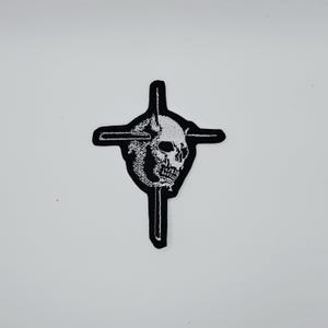 Puede incluir: Parche bordado en blanco y negro con un cráneo dentro de una cruz. El cráneo tiene detalles. El parche tiene un borde cosido, diseñado para su aplicación en ropa o accesorios. Ideal para la moda alternativa.