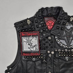 K&ouml;nnte beinhalten: Schwarze Lederweste mit Metallnieten und Aufn&auml;hern. Die Weste zeigt einen "Hellhammer" Aufn&auml;her mit einer d&auml;monischen Illustration und dem Text "Demon Entrails". Weitere Aufn&auml;her sind ein Pentagramm und ein Ziegenkopf-Design. Sch&auml;del-f&ouml;rmige Anstecknadeln sind ebenfalls vorhanden.