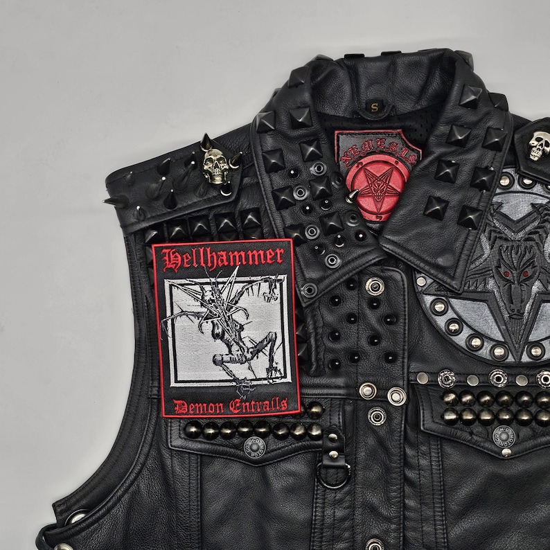 K&ouml;nnte beinhalten: Schwarze Lederweste mit Metallnieten und Aufn&auml;hern. Die Weste zeigt einen "Hellhammer" Aufn&auml;her mit einer d&auml;monischen Illustration und dem Text "Demon Entrails". Weitere Aufn&auml;her sind ein Pentagramm und ein Ziegenkopf-Design. Sch&auml;del-f&ouml;rmige Anstecknadeln sind ebenfalls vorhanden.
