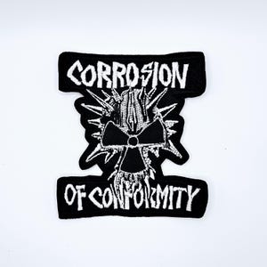 May include: Black and white embroidered patch with the words "CORROSION" and "OF CONFORMITY". The patch features a central design of a stylized radiation symbol with spiky elements.