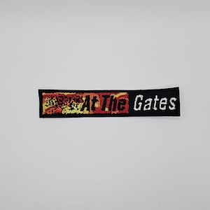 Pode incluir: Patch retangular preto com o texto "At The Gates" em letras brancas e vermelhas. O canto superior esquerdo tem um design desgastado em tons de laranja, amarelo e vermelho.