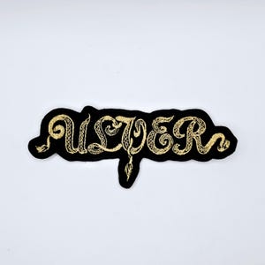 May include: Black embroidered patch with the word "ULVER" in gold thread. The letters are stylized with a snake-like design. The patch has a black border and is set against a white background.