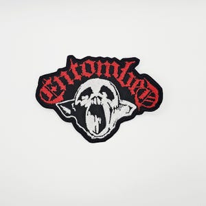May include: Embroidered patch with the word "Entombed" in red, stylized lettering. The patch features a black background with a white skull graphic. The skull has an open mouth and is surrounded by a black border.