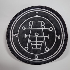May include: A black and white embroidered patch with a circle design and a symbol inside. The symbol is a white design on a black background. The circle is surrounded by a white border.