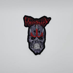 Op de afbeelding: Geborduurde patch met een schedelontwerp, rode accenten en het woord "MONSTROFIED" in rood. De schedel heeft een kroonachtig ontwerp erboven en is zwart omlijnd.