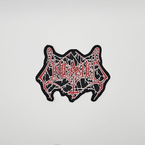 May include: Black and red embroidered patch with the word "UNLEASHED" in a gothic font. The patch has a spiderweb design and a cross symbol. The patch is on a white background.