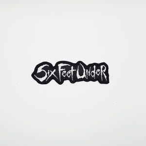 Puede incluir: Un parche bordado en blanco y negro con el texto "Six Feet Under" en una fuente gótica. El parche tiene un borde negro y está sobre un fondo blanco. El texto es blanco.