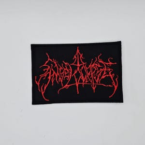 Puede incluir: Parche rectangular negro con texto rojo bordado. El texto está estilizado y parece ser un logotipo o emblema de una banda. El parche es probablemente para ropa o accesorios.