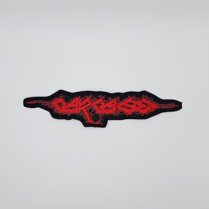 May include: A black embroidered patch featuring red text. The text is in a stylized font. The patch has a distinctive shape, with a black border framing the red text.