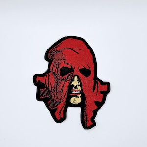 Könnte beinhalten: Gestickter Aufnäher mit einer roten Kapuzenfigur mit schwarzen Augenlöchern und einem teilweise sichtbaren Gesicht. Der Aufnäher hat einen schwarzen Rand und ist vor einem weißen Hintergrund platziert. Das Design ist detailliert und scheint maschinell gestickt zu sein.