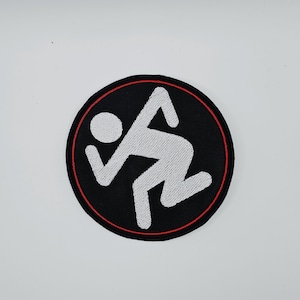 Op de afbeelding: Geborduurde patch met een witte, gestileerde rennende figuur op een zwarte cirkelvormige achtergrond. Een dunne rode rand omringt de zwarte cirkel. Geschikt voor kleding of accessoires.