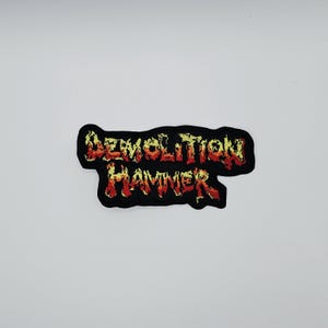 May include: A black embroidered patch with the words "DEMOLITION HAMMER" in red and yellow flame-like lettering. The patch has a black border and is set against a white background.