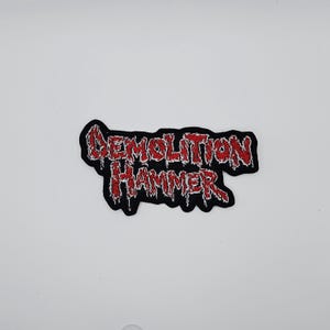 Może przedstawiać: Haftowana naszywka z napisem "DEMOLITION HAMMER" w kolorze czerwonym, obramowana czarnym kolorem na czarnym tle. Naszywka przeznaczona jest do umieszczenia na odzieży lub akcesoriach.
