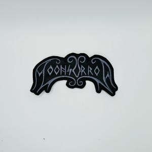 May include: An embroidered patch featuring a black background with light grey stitching. The design displays stylised, angular lettering that reads MOONHORROR, framed by decorative, symmetrical scrollwork elements at the top and bottom edges.