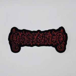 May include: Black embroidered patch with the word "DISSECTION" in red, stylized lettering. The patch has a flame-like design around the edges and is rectangular with rounded ends. The patch is likely for clothing or accessories.