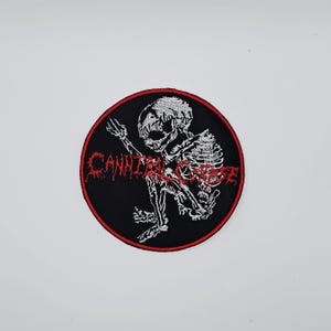 May include: A circular black patch with a red trim, displaying a white skeleton design. The red text reads "CANNIBAL CORPSE". This patch is designed for application on clothing or accessories.