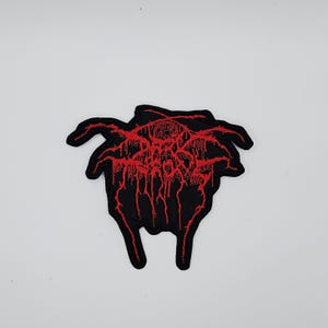 May include: Black embroidered patch with a red, stylized design. The patch features a complex, abstract pattern with a dripping effect. The patch has a unique shape with two downward-pointing extensions.