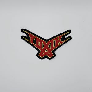 Pode incluir: Patch bordado com a palavra "TOXIR" em vermelho, contornado em dourado, sobre fundo preto. O patch tem um design angular único, que se assemelha a um pássaro ou emblema estilizado. Ideal para roupas ou acessórios.
