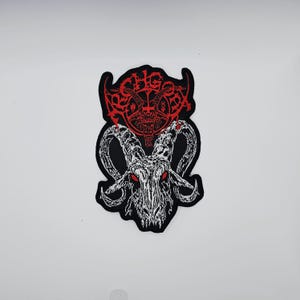 May include: Embroidered patch featuring a white goat skull with red eyes and large curved horns. Above the skull is a red emblem with intricate lettering and a pentagram. The patch has a black border and is likely for clothing or accessories.