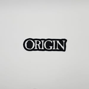 Puede incluir: Un parche bordado en blanco y negro con la palabra "ORIGIN". El texto es blanco, en negrita, sin serifa, con un borde negro. El parche rectangular está sobre un fondo blanco.