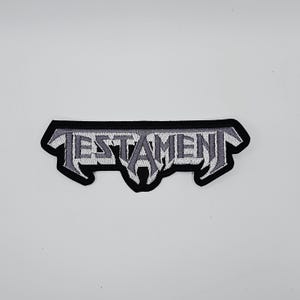 May include: Embroidered patch featuring the word "TESTAMENT" in a stylized, gothic font. The text is in shades of gray and white, outlined in black, and set against a black background. The patch has a unique, angular shape.