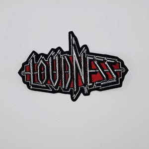 Puede incluir: Parche bordado con la palabra "LOUDNESS" en blanco, contorneada en negro, sobre un fondo rojo. El parche tiene un diseño estilizado y dentado, con un borde negro. Ideal para chaquetas, bolsos u otros artículos de tela.